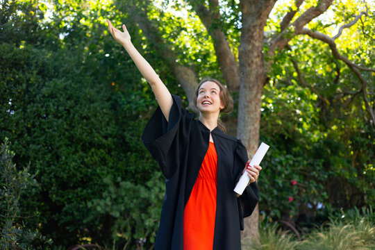 Woman standing in garden by tree trunk, wearing gown, holding diploma, raising arm and smiling