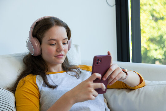 Adult woman reclining on sofa at home in mustard raglan shirt, using pink smartphone and headphones