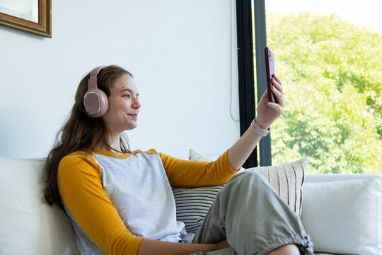 Adult female sitting on white couch near window wearing pink headphones and holding red smartphone