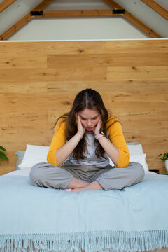 Woman sitting cross-legged on loft bed with wooden headboard, wearing yellow top, cupping cheeks