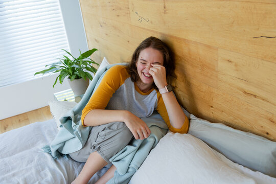 Adult woman laughing, covering face, sitting on bed, wrapping in teal blanket, smartwatch, plant