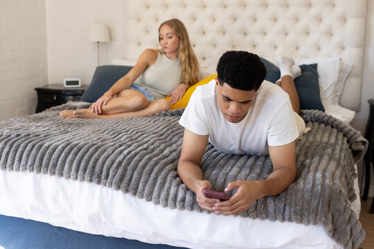 Couple sharing bed, man lying on stomach holding smartphone, woman reclining near tufted headboard