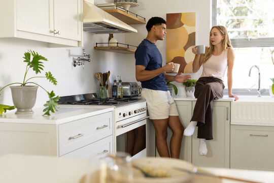 Diverse couple chatting and holding ceramic mugs in bright home kitchen, wearing casual clothes