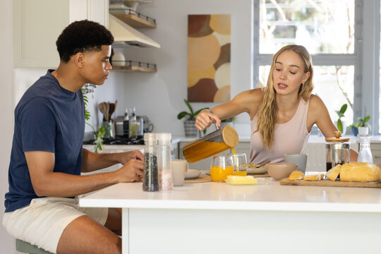 Diverse couple sitting at white kitchen island, woman pouring juice from glass pitcher