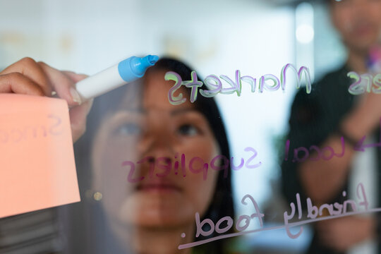 Diverse coworkers collaborating in office at glass board, holding blue marker, wearing blazer