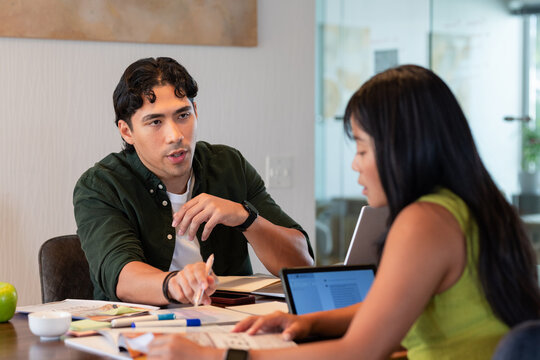 Diverse coworkers wearing green shirt and olive top, reviewing documents on laptop at meeting table