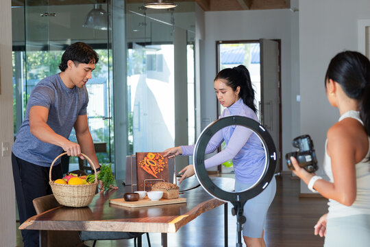 Diverse team arranging carrot cookbook and fruit basket on liveedge table, filming DSLR ring-light