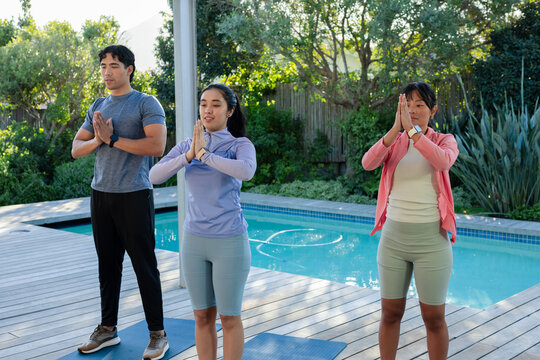 Diverse friends practicing yoga on wooden pool deck beside pool, wearing athletic wear, blue mats