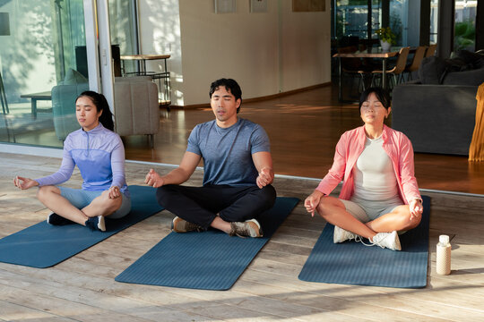 Diverse friends in sportswear meditating on wooden deck on mats near glass doors with water bottle
