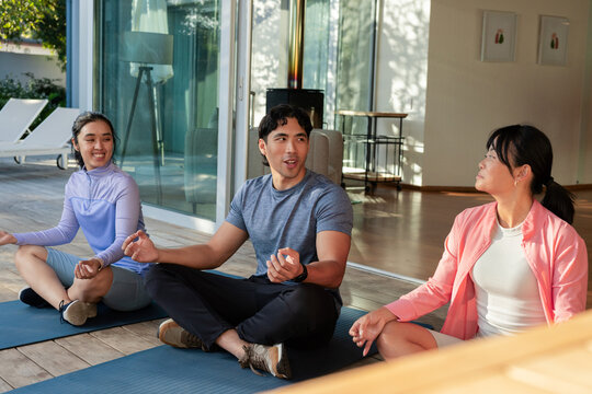 Diverse friends sitting cross-legged on blue mats on deck by glass doors wearing sportswear