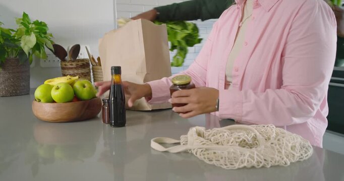 Green-jacket person is pulling celery from paper bag while woman is moving jars, restocking kitchen