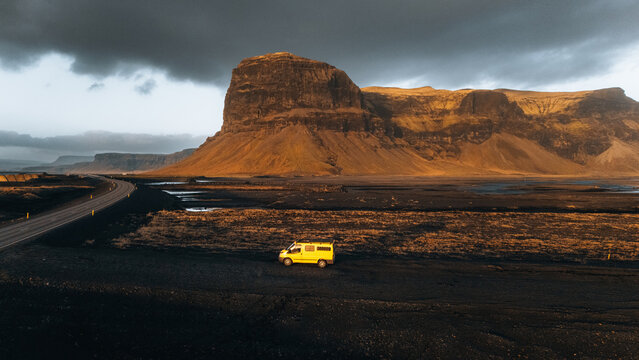 Aerial view of the Lomagnupur mountain with a yellow van parked on the dark volcanic plains under a dramatic sky in Skaftarhreppur, Iceland.