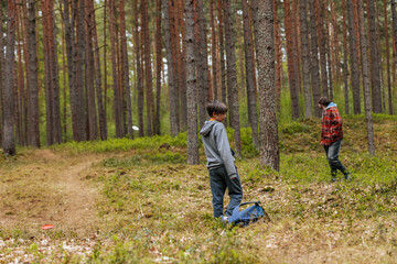 Fototapeta premium Two boys playing disc golf in a forested area, one boy wearing a gray hoodie and the other in a red plaid jacket, surrounded by tall pine trees and green underbrush