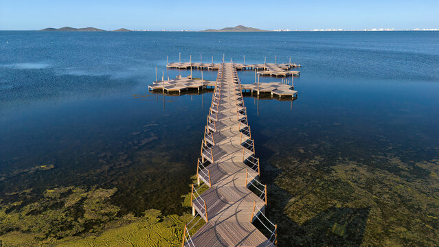 Aerial view of a wooden pier and bathing platforms over calm shallow water in Los Urrutias, on the Mar Menor lagoon in Murcia, Spain, with distant islands and blue sea.