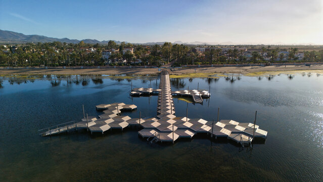 Aerial view of a wooden bathing pier facing the beach, palm trees and coastal houses in Los Urrutias, beside the calm Mar Menor lagoon in Murcia, Spain.