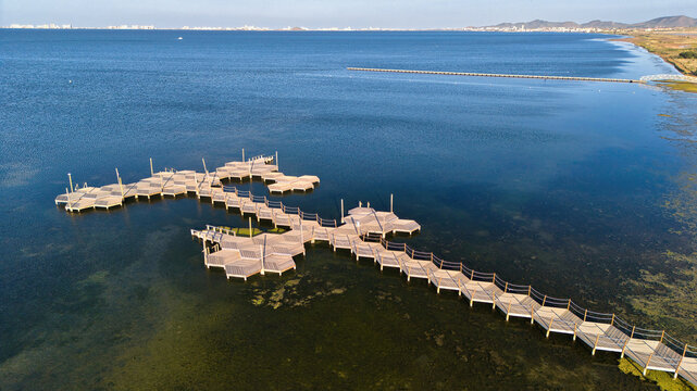 Aerial landscape of wooden bathing platforms and a curved pier over shallow blue water in Los Urrutias, Murcia, with the Mar Menor coastline and distant mountains.