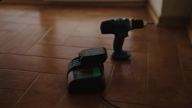 Lithium ion battery pack for cordless electric drill charging in power station on wooden floor of room, with hand tool waiting in background for house renovation, no people, nobody, slow motion.