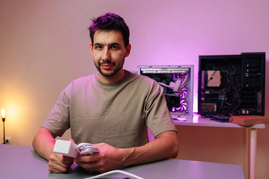 Confident technician holding power adapter at desk. Open PC cases in background with purple neon light. IT support, hardware repair, custom computer building, maintenance service concept.