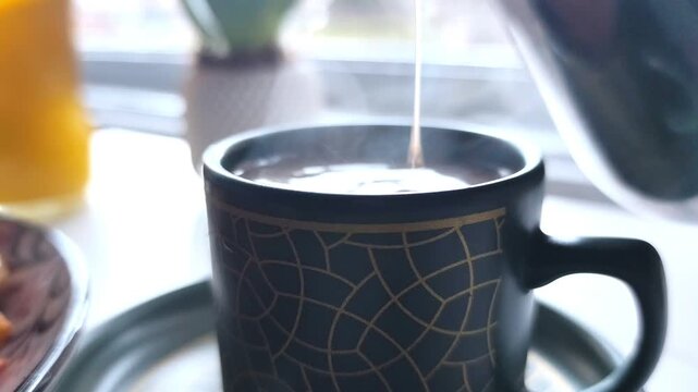 Static close-up shot of hot coffee being poured from a silver pitcher into a dark green patterned ceramic cup on a white table.