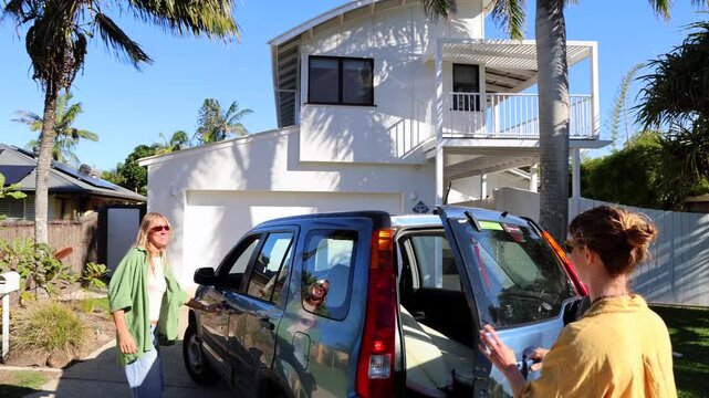 Two female surfers talking and loading a surfboard into the trunk of their car, preparing for a road trip to the beach on a sunny summer day in a suburban neighborhood with palm trees