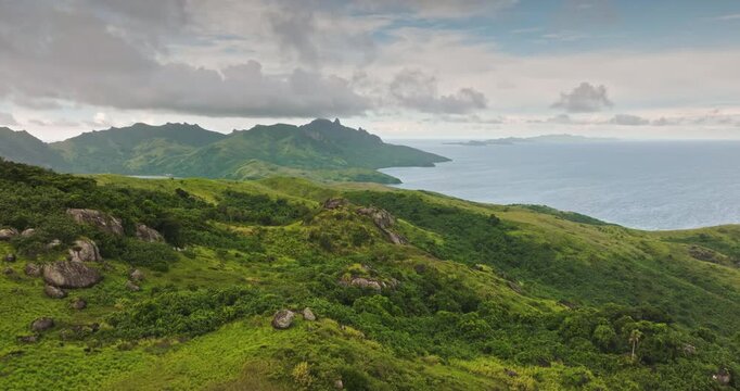 Fiji island landscape with lush tropical mountains, rolling green hills, volcanic peaks meeting clear Pacific Ocean under cloudy sky, natural beauty and remote environment. Aerial view drone flight