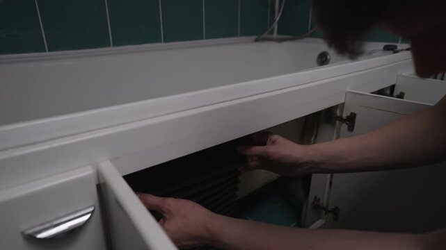 Male hands placing mysterious black object inside hidden storage space under bathtub, concealing it from view in bathroom setting with green tiled walls.
