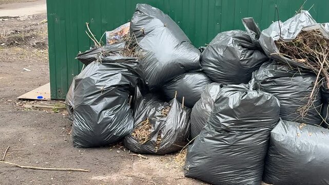 Black garbage bags piled by fence, on roadside near small green trash can, scattered leaves and gravel, neglected urban corner