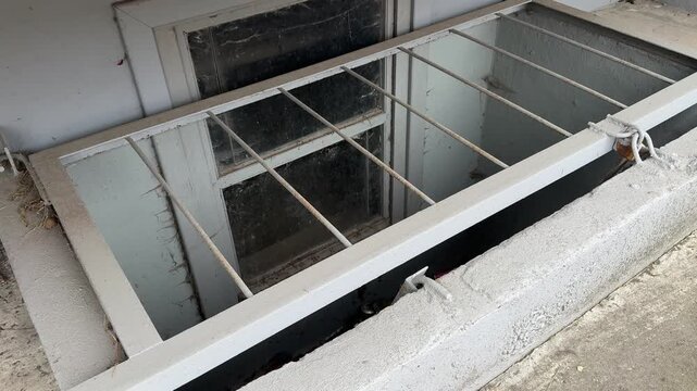 Metal grating on the basement window, concrete ledge under the balcony of a high-rise building, spring daylight, weathered paint, moss on the asphalt, chips
