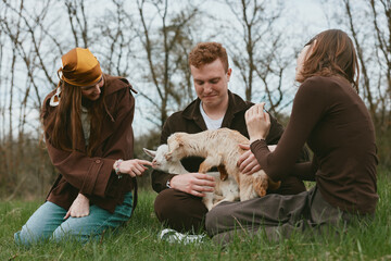 Obraz premium Three people share a playful moment with a goat kid in a grassy meadow, outdoors lifestyle scene. They laugh and interact, showcasing bonding, care, and rustic countryside vibes in natural light.