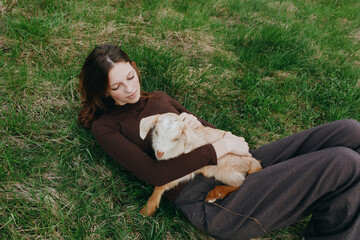 Obraz premium Outdoor scene of a woman lying on lush grass, gently cuddling a goat kid, sun warmed, cozy and relaxed, showcasing lifestyle, companionship, and a deep connection with nature