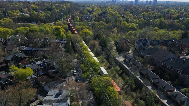 Freight train with intermodal containers moving through a residential neighborhood in Canada, during a sunny spring day showing green trees and suburban houses