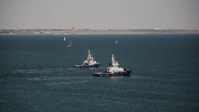 Two tugboats move across calm blue water near coast of Odessa Ukraine. Scene shows industrial maritime transport with clear visibility and seascape featuring distant shoreline and small boats