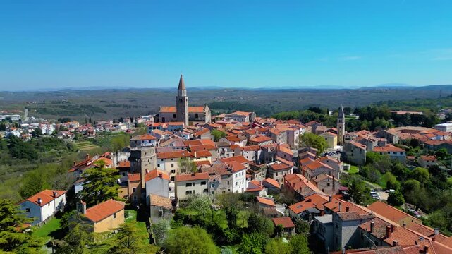 Aerial view of Buje, a charming hilltop village in Istria, Croatia, bathed in bright midday sun. Ancient architecture and lush spring greenery define this picturesque destination.
