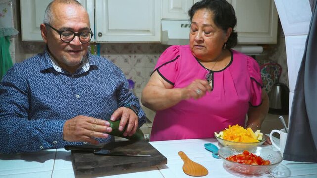 An elderly woman shows her husband how to peel and slice a cucumber on a cutting board