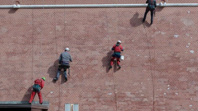 Three Workers Inspecting Red Brick Wall, Safety Supervisor Leads Routine Inspection With Technicians Descending On Ropes, Checking Anchors And Mortar,