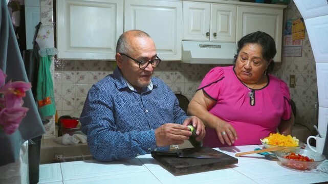 An elderly woman shows her husband how to peel and slice a cucumber on a cutting board