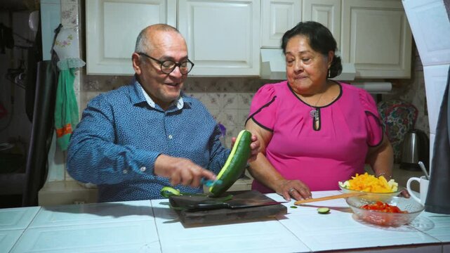 An elderly woman shows her husband how to peel and slice a cucumber on a cutting board
