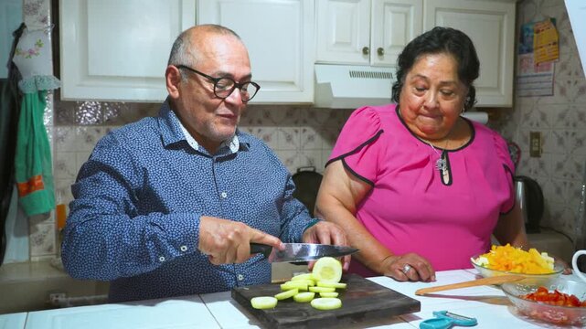 An elderly woman shows her husband how to peel and slice a cucumber on a cutting board