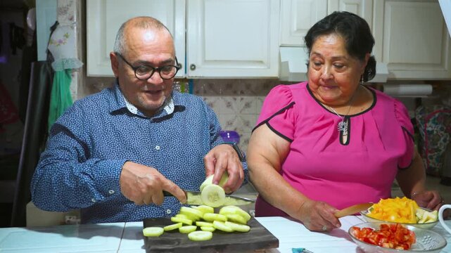An elderly woman shows her husband how to peel and slice a cucumber on a cutting board