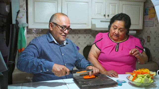 An elderly woman shows her husband how to peel and cut a carrot on a cutting board
