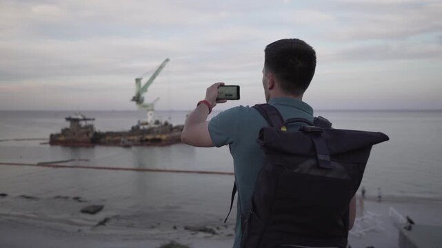 Male blogger is broadcasting live on social networks on journey near sunken tanker Delfi near Odessa. Black Sea coast. Man creating video blog content using smartphone camera about ship accident.