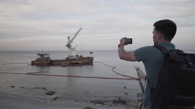 Male blogger is broadcasting live on social networks on journey near sunken tanker Delfi near Odessa. Black Sea coast. Man creating video blog content using smartphone camera about ship accident.