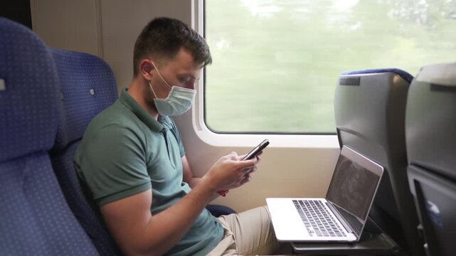 Theme is work on a trip. Young Caucasian male freelancer texting on a smartphone and working on a laptop wearing a protective medical mask on his face while sitting in a train carriage by the window.