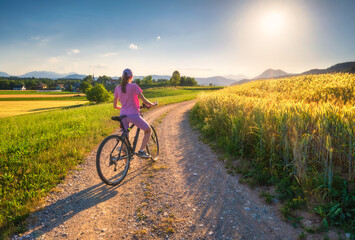 Fototapeta premium Woman on mountain bike on gravel road at sunset in summer. Colorful landscape with sporty girl riding bicycle, blooming fields, meadows with yellow and green grass, dirt road, sky. Sport and travel