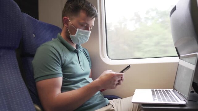 Man working in train, wearing masks to prevent air pollution, dust, PM2.5 pm2, coronavirus or COVID 19. Male passenger is surfing internet on smart phone and uses laptop near window. Railway transport