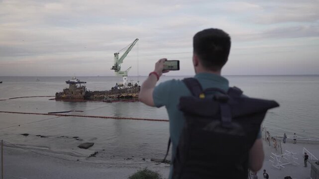 Young traveler blogger takes selfie on phone, makes video broadcast for subscribers on Sea coast about sunken tanker Delf, its rescue using floating crane. Shooting vlog, streaming online podcast.