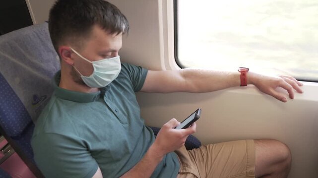 Public transport, man traveling inside train sitting keeping safety distance and mask, sitting using the phone. Male use smart phone inside train car. Traveler wears mask to prevent spread Covid 19.