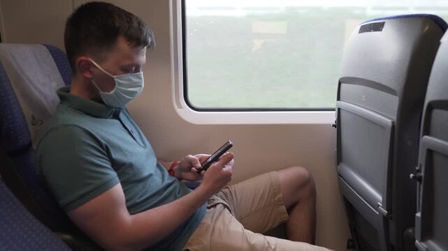 Man using mobile phone on a high speed train. Male on mask face protection, use smartphone and keep social distancing while commuting in public transport. Travelling by train with coronavirus.