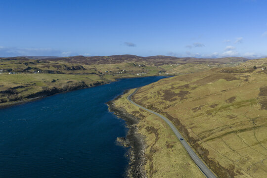 Aerial view of Gesto Bay with a winding coastal road and rolling hills under a clear blue sky in Isle of Skye, Scotland, United Kingdom.
