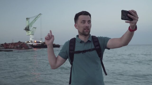 Young traveler blogger takes selfie on phone, makes video broadcast for subscribers on Sea coast about sunken tanker Delf, its rescue using floating crane. Shooting vlog, streaming online podcast.